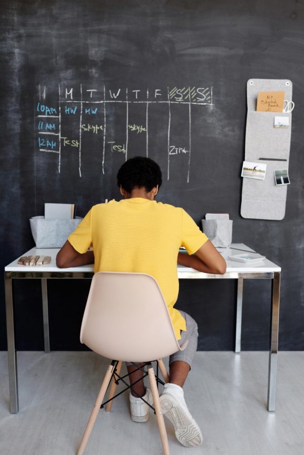 Boy sits working at desk in front of chalkboard with weekly schedule written on it.